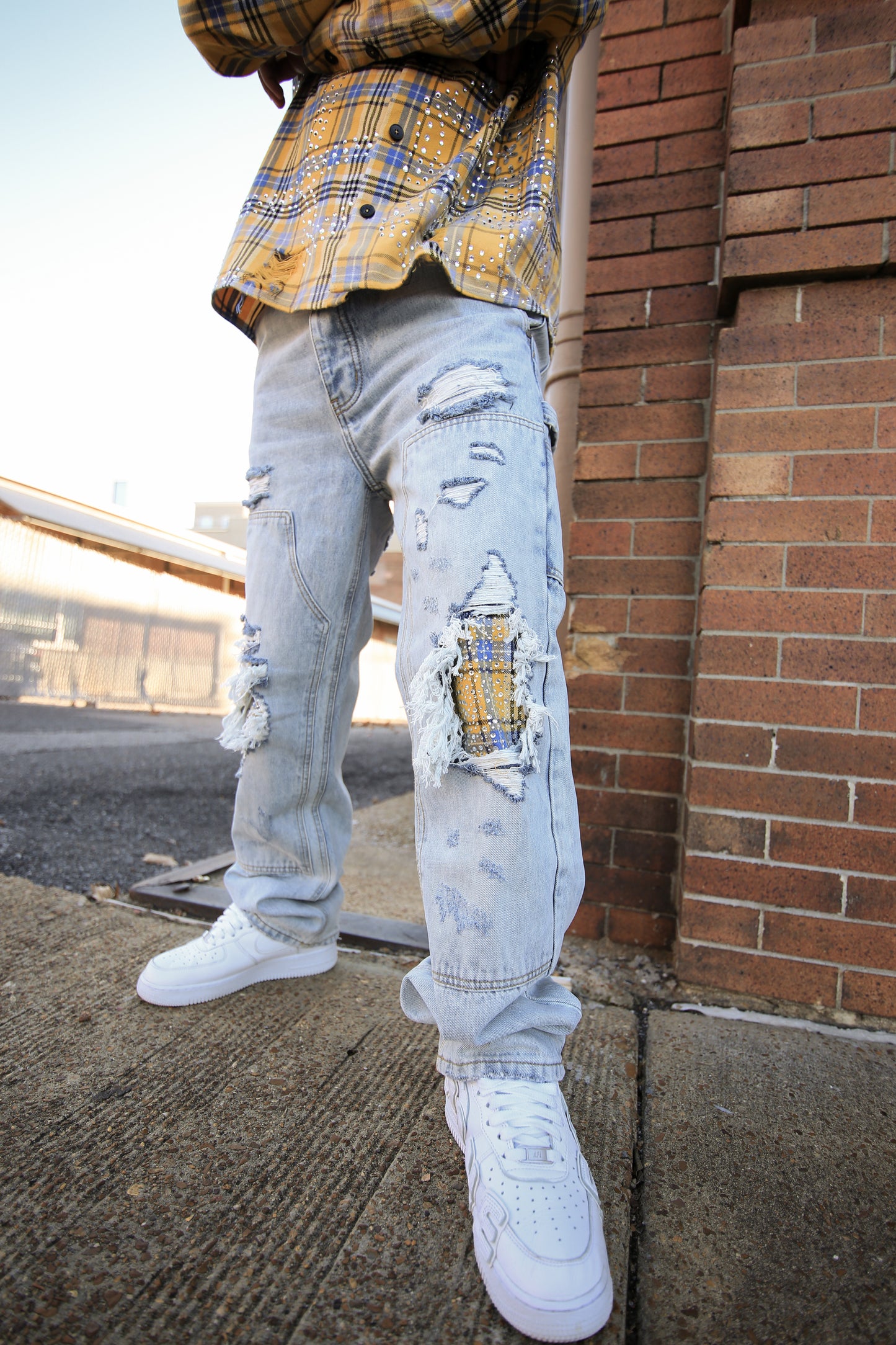 Person wearing a yellow plaid shirt, light blue ripped jeans, and white sneakers against a brick wall.