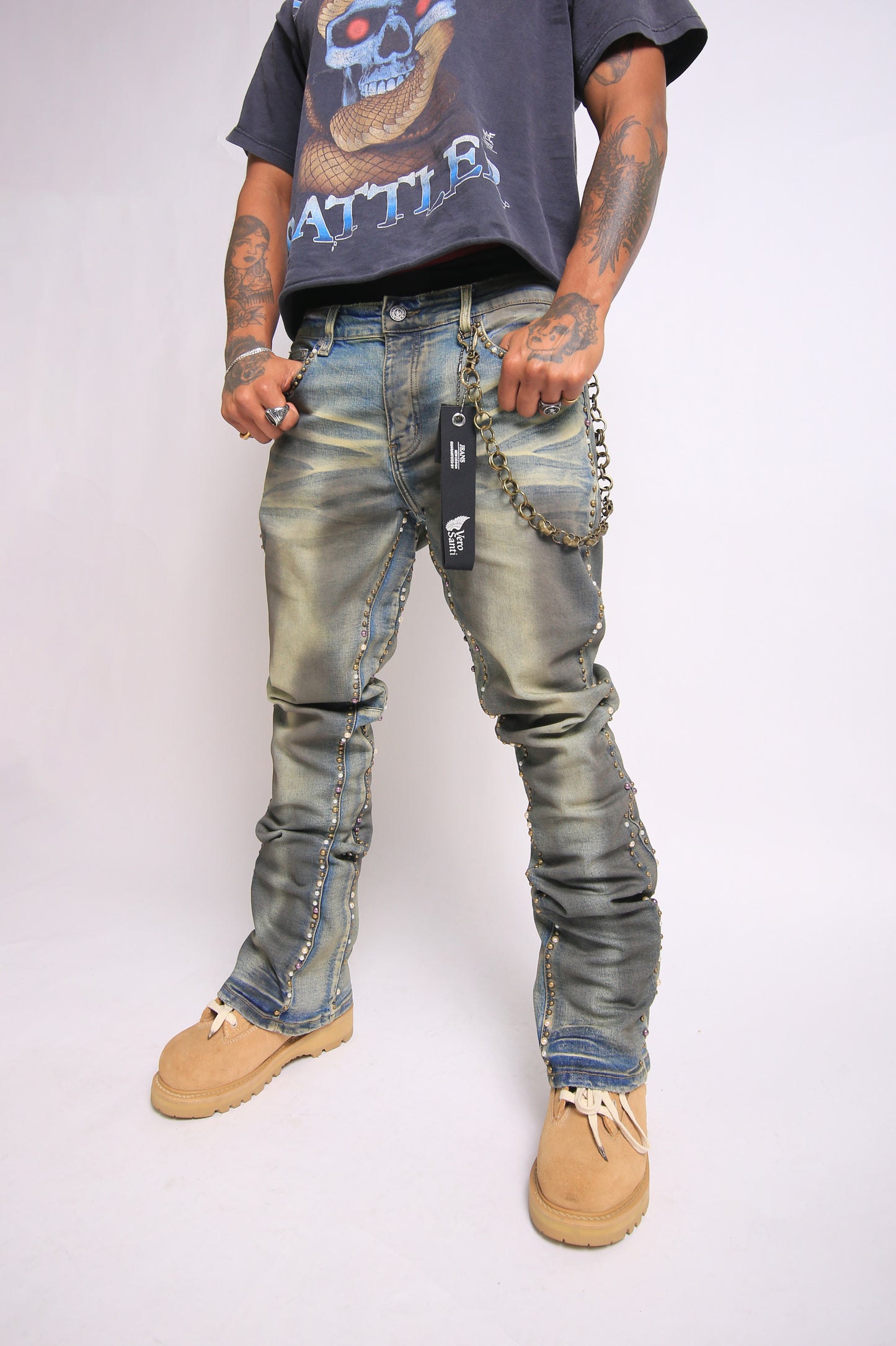 Person wearing distressed jeans with patches and a chain, standing against a plain background