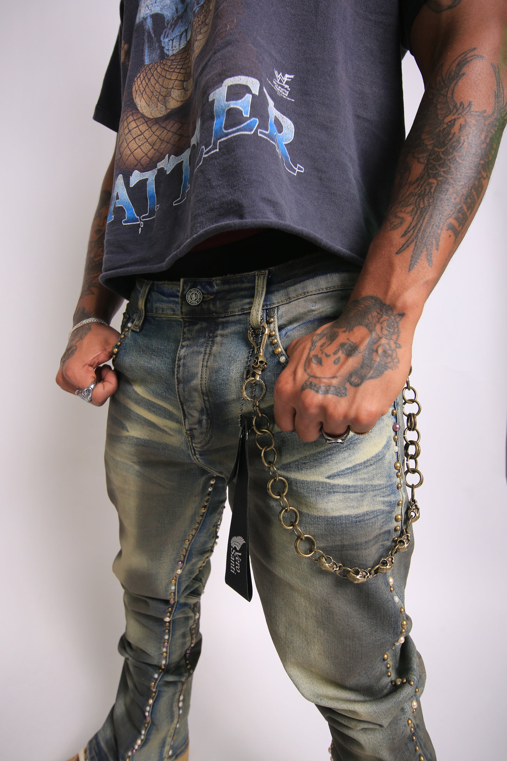 Person wearing a dark t-shirt with visible branding and distressed jeans with chains, on a plain background.