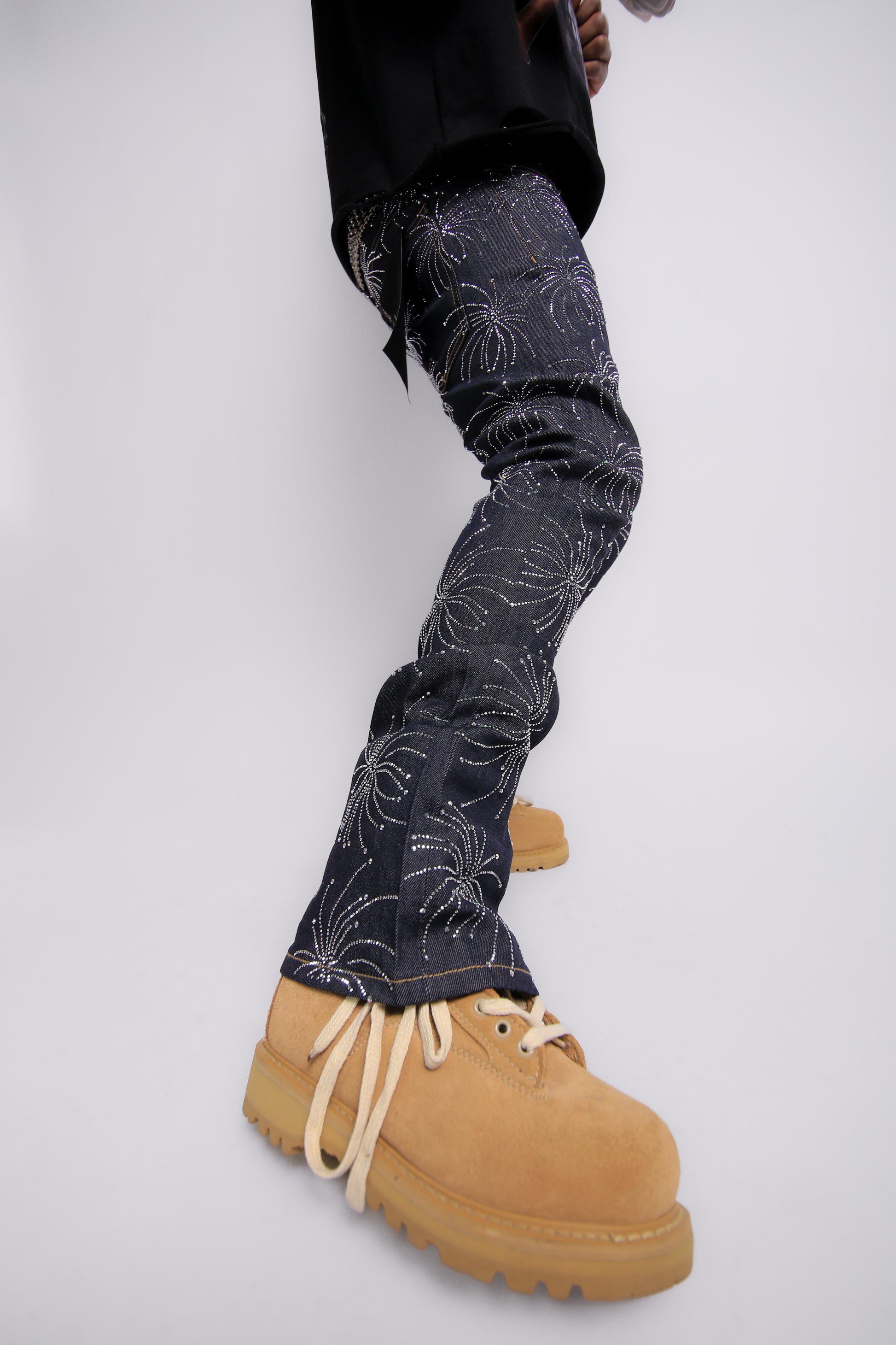 Person wearing dark jeans with floral patterns and tan boots on a light gray background